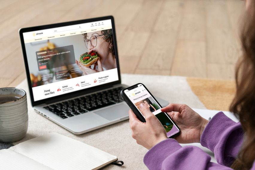 Person using a laptop and smartphone to browse a health-related website, sitting with a cup of coffee and notebook nearby.