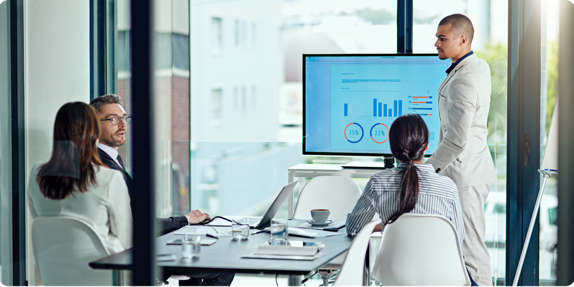 Business meeting in a glass-walled office with a man in a suit presenting financial charts on a screen to seated colleagues.