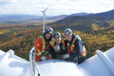 Drei Handwerker in voller Klettermontur hoch oben auf einer Windkraftanlage schauen lächelnd in die Kamera. Im Hintergrund sanfte Hügel und ein weiter Blick über Herbstwälder.