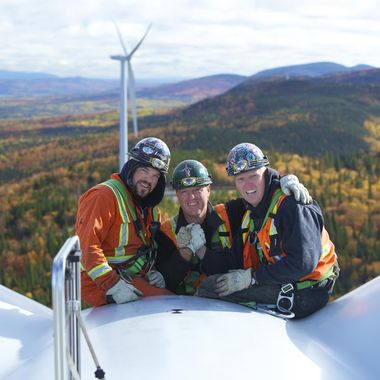 Handwerker sitzen auf einem Windrad weit oben über einer hügeligen Waldfläche.