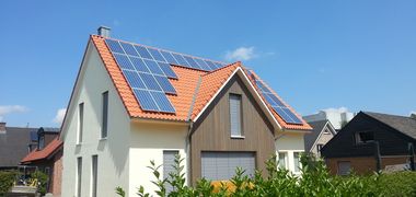 Solar Paneele auf dem roten Ziegeldach eines Einfamilienhauses, das Haus ist eingerahmt von einer grünen Hecke und blauem Himmel.