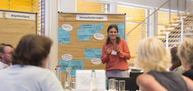 Eine Frau erzählt der Kleingruppe eines Bürgerrates etwas vor einer Wand mit handgeschriebenen Kärtchen.