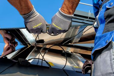 Zwei Menschen heben mit Arbeitshandschuhen eine silberne, verkabelte Platte in eine Fassung.