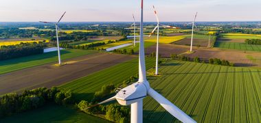 Mehrere Windräder aus der Vogelperspektive inmitten von grünen Feldern und Abendsonne.