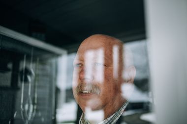 Ein älterer Mann steht hinter einer Glasscheibe und schaut freundlich heraus.