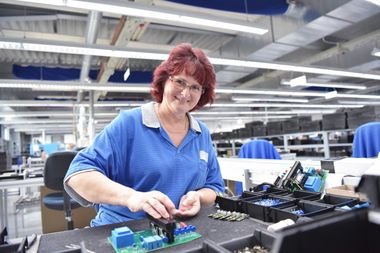 Eine Frau sitzt in einer großen, hellen Werkshalle. Auf ihrem Tisch liegen elektronische Bauteile und eine Platine.
