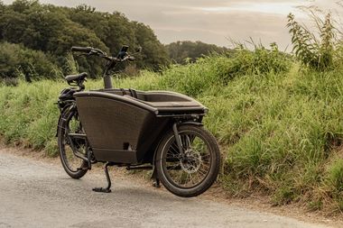 Ein schwarzes Lastenrad auf eine Straße neben Wiese und Wäldern.