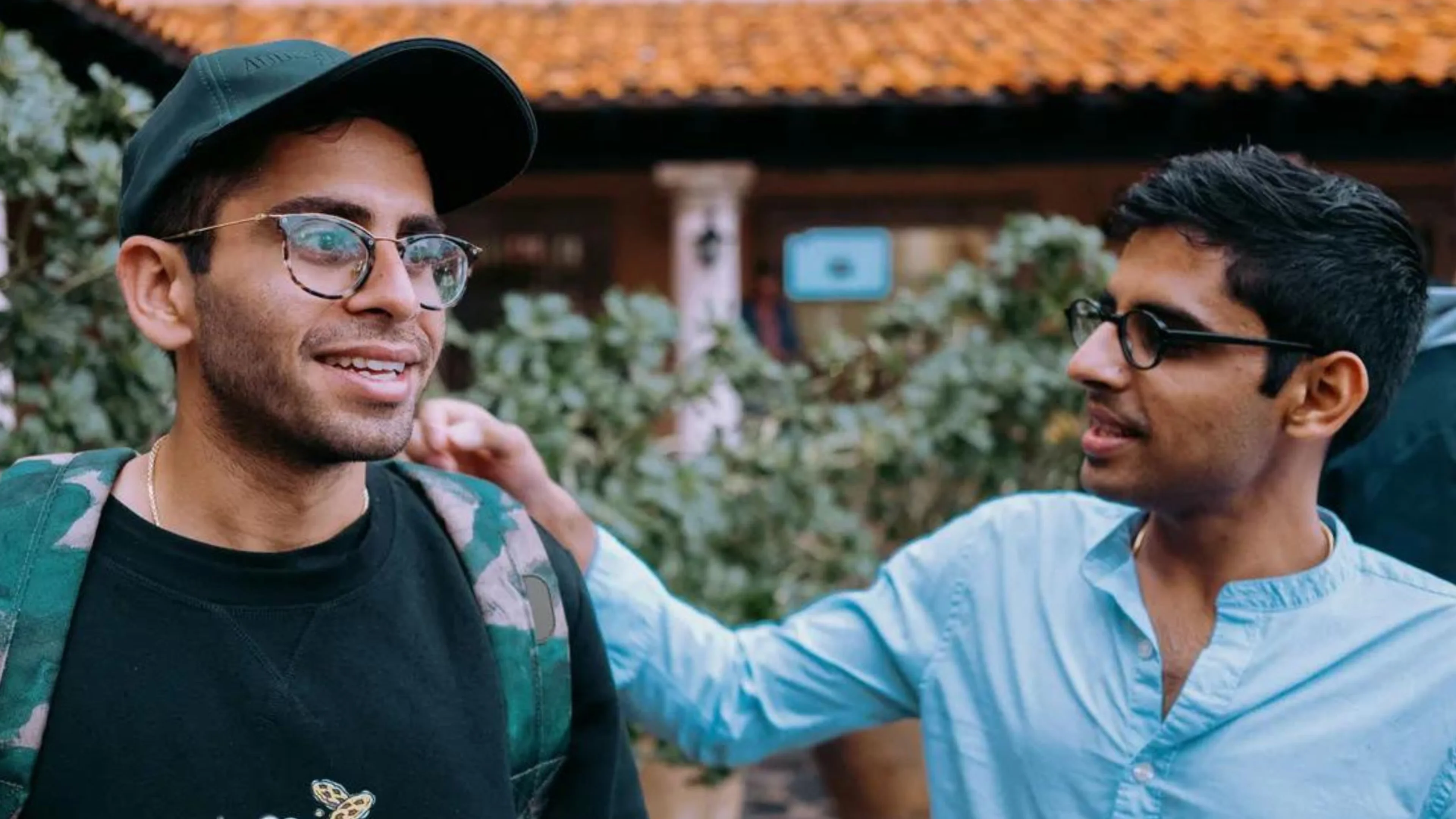 Two slightly smiling  men standing beside's each other, outdoors, one has hand on the other's shoulder in a friendly manner.