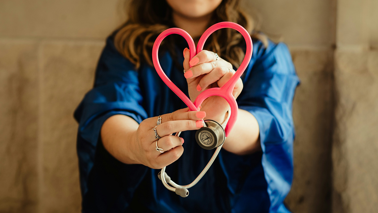 A person wearing a blue outfit holds a pink stethoscope shaped into a heart.