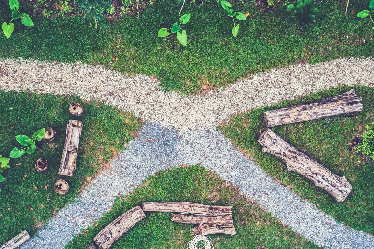 Aerial view of a garden with intersecting gravel paths, surrounded by green grass and wooden logs arranged decoratively.