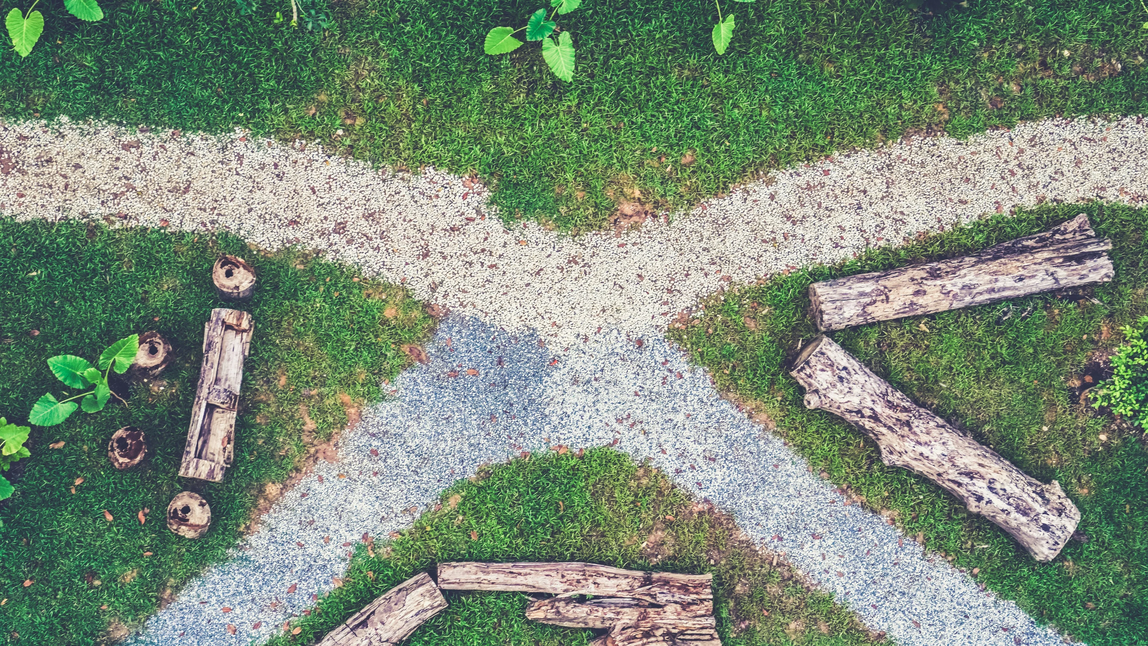 Aerial view of a garden with intersecting gravel paths, surrounded by green grass and wooden logs arranged decoratively.