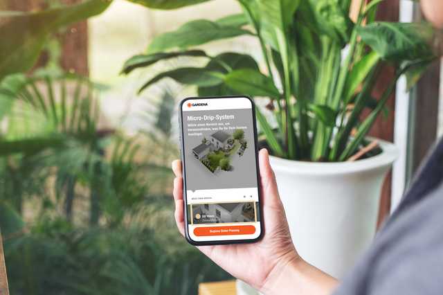 Hand-held smartphone showing a micro-drip garden app, person seated by a wooden table with large potted plants in the background.
