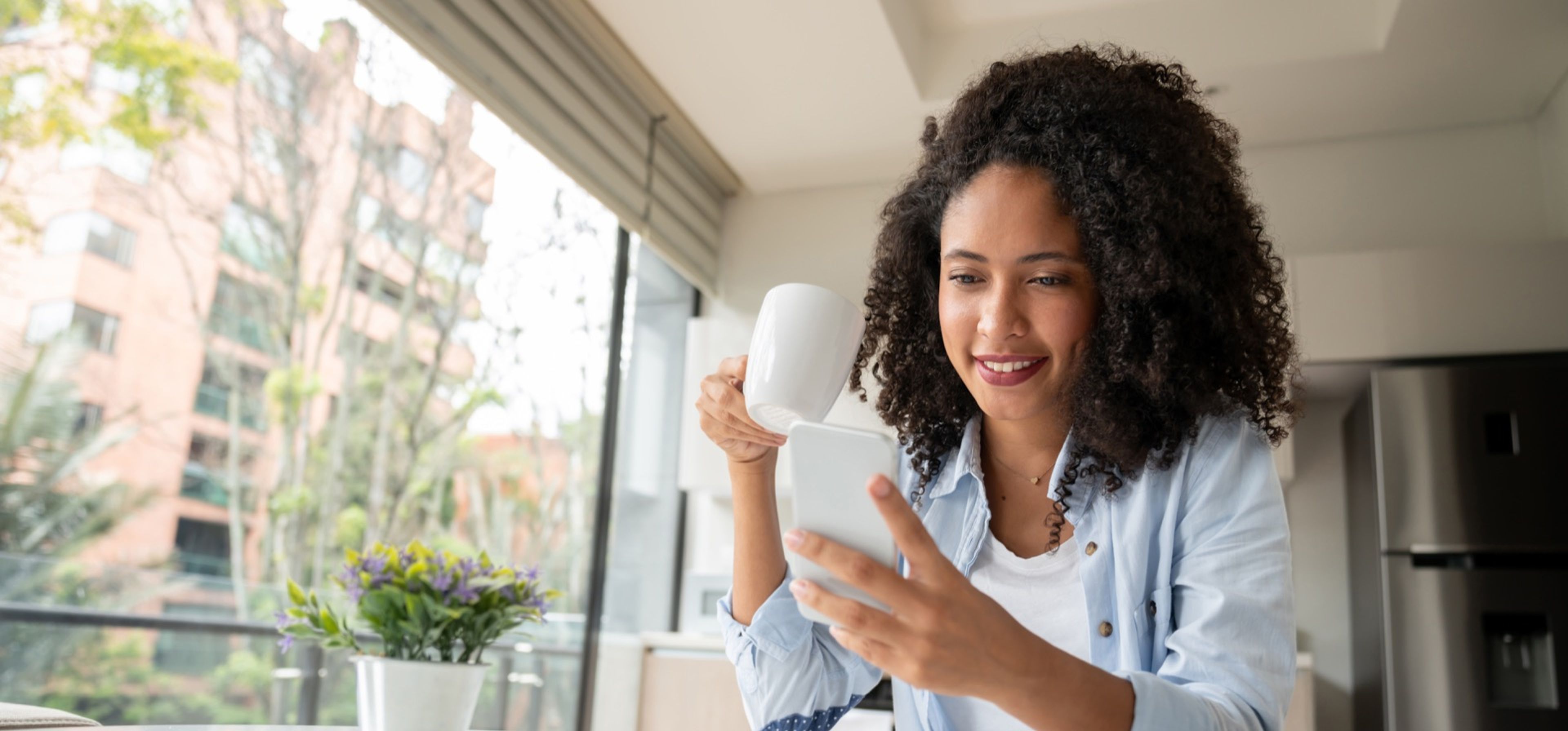 Eine Frau mit Smartphone und Kaffeetasse am Küchentisch.