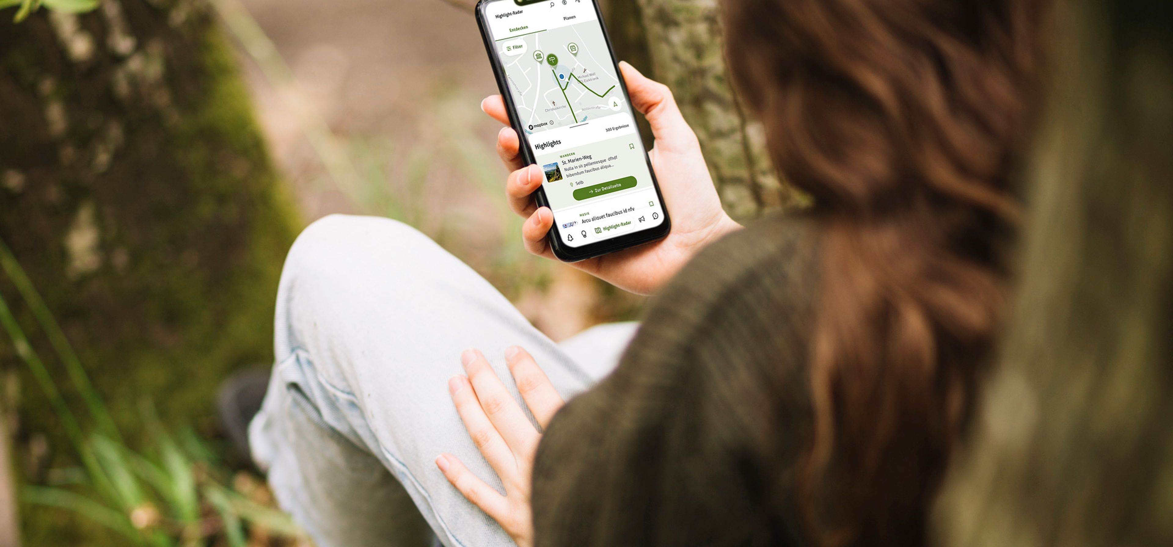 Person hält ein Smartphone mit einer Wander-App in der Hand, während sie im Wald sitzt.
