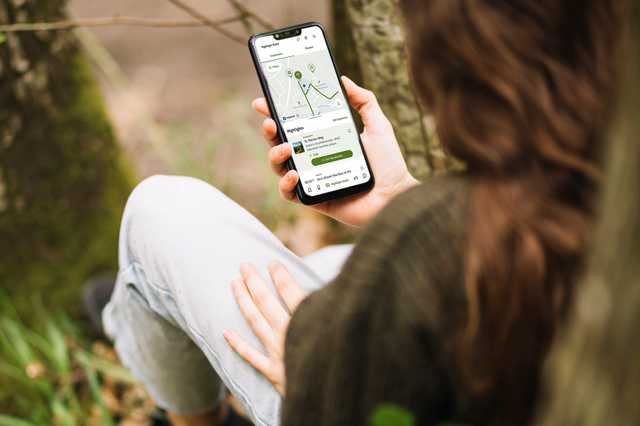 Person hält ein Smartphone mit einer Wander-App in der Hand, während sie im Wald sitzt.