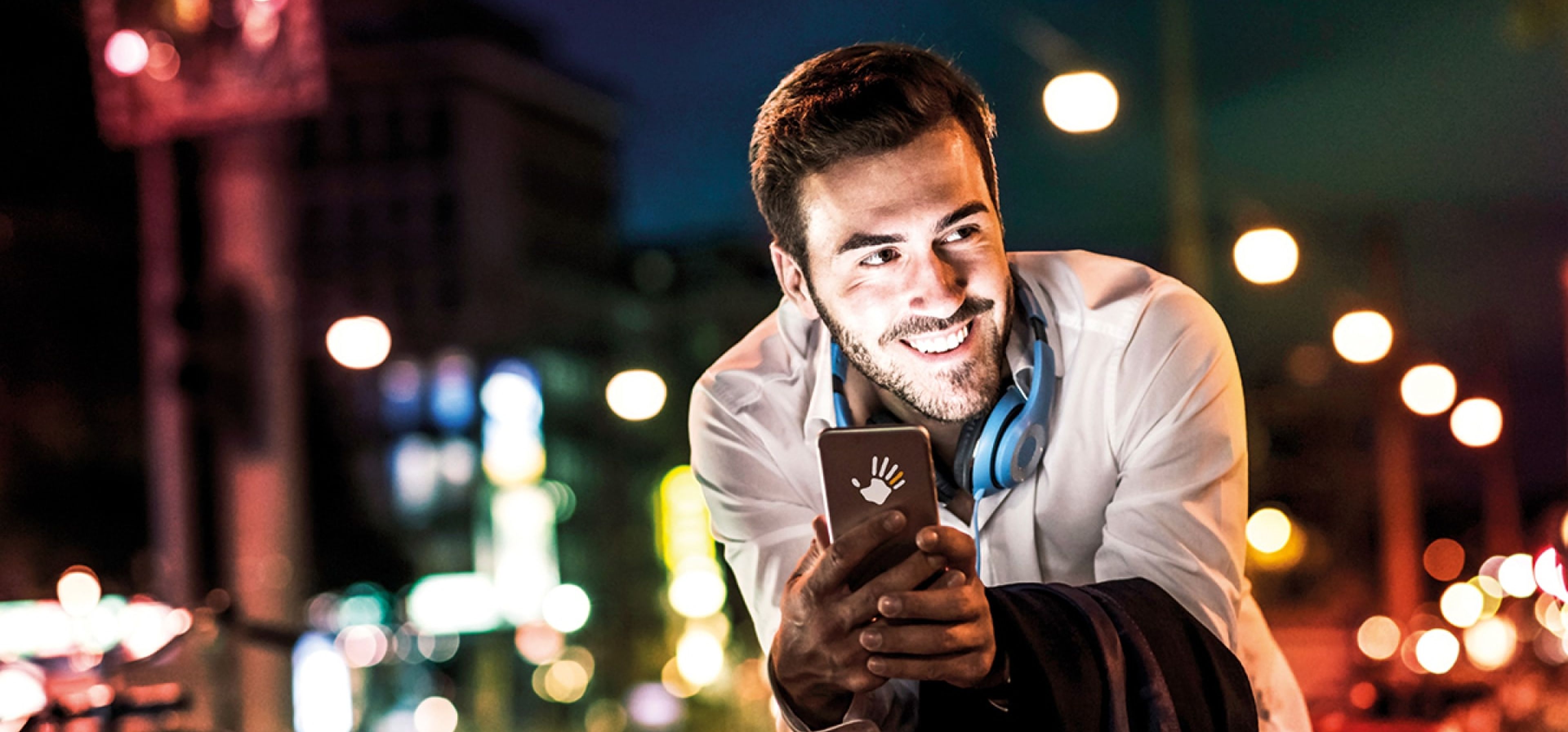 Lächelnder Mann mit Smartphone in der Hand lehnt an seinem Fahrrad in einer Stadt bei Nacht.