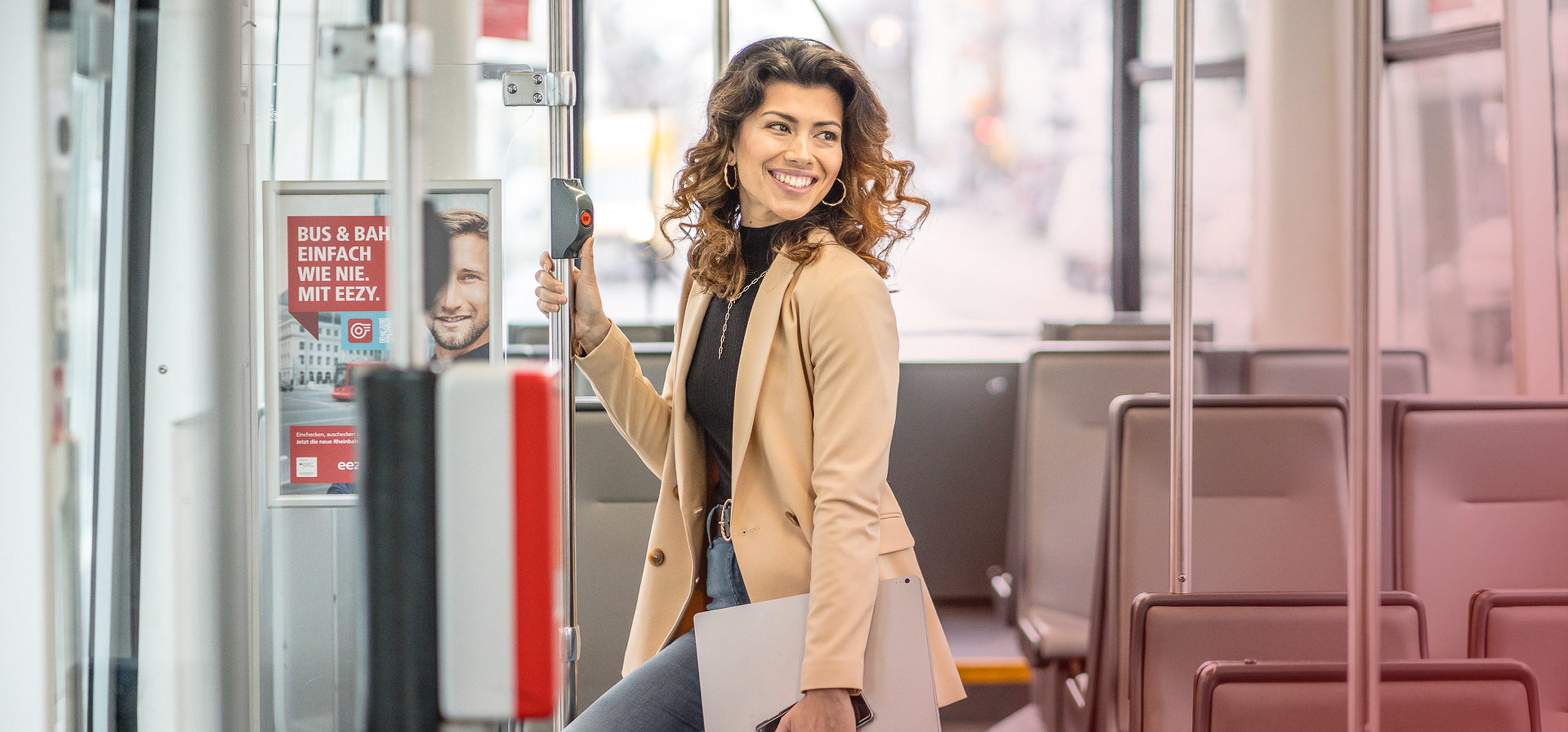 Frau steht in einer Straßenbahn mit ihrem Lapto. Sie lächelt und hält sich fest.