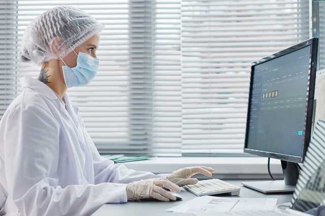 Wissenschaftliche Fachkraft mit Haarnetz, Maske und Handschuhen arbeitet am Computer
