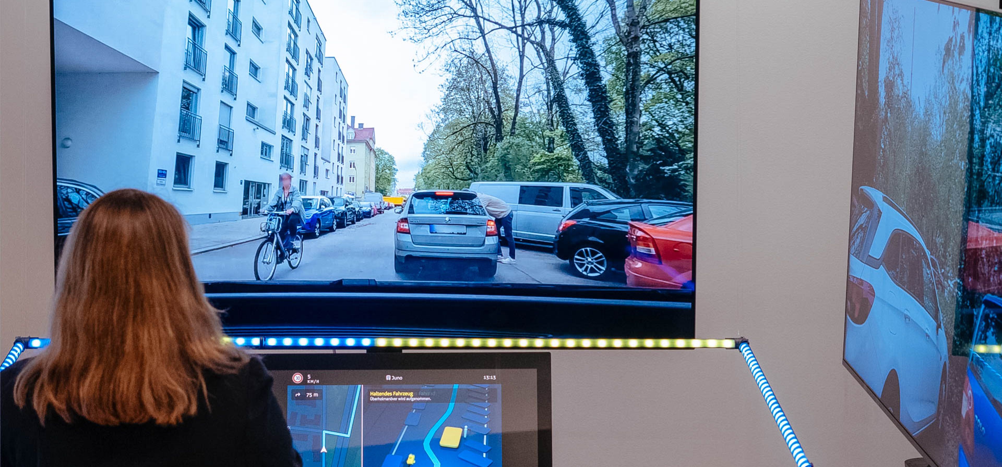 A person interacts with a driving simulator, displaying a city street with cars and a cyclist. A digital map is visible on the control panel.
