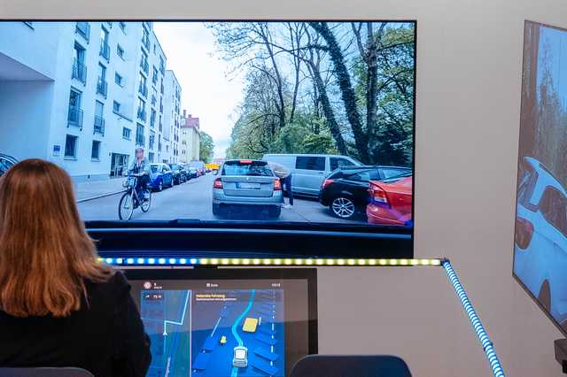 A person interacts with a driving simulator, displaying a city street with cars and a cyclist. A digital map is visible on the control panel.