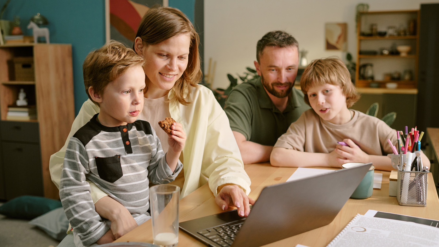 A family of four sits at a table, smiling and looking at a laptop. The mother holds a child eating a cookie, while the father and another child watch.