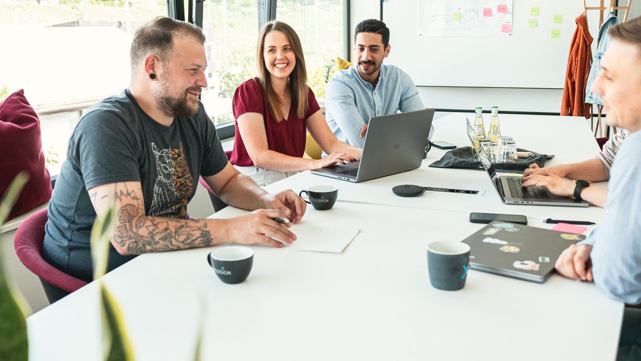 Vier Personen sitzen lächelnd um einen Tisch in einem modernen Büro, arbeiten an Laptops und diskutieren angeregt.