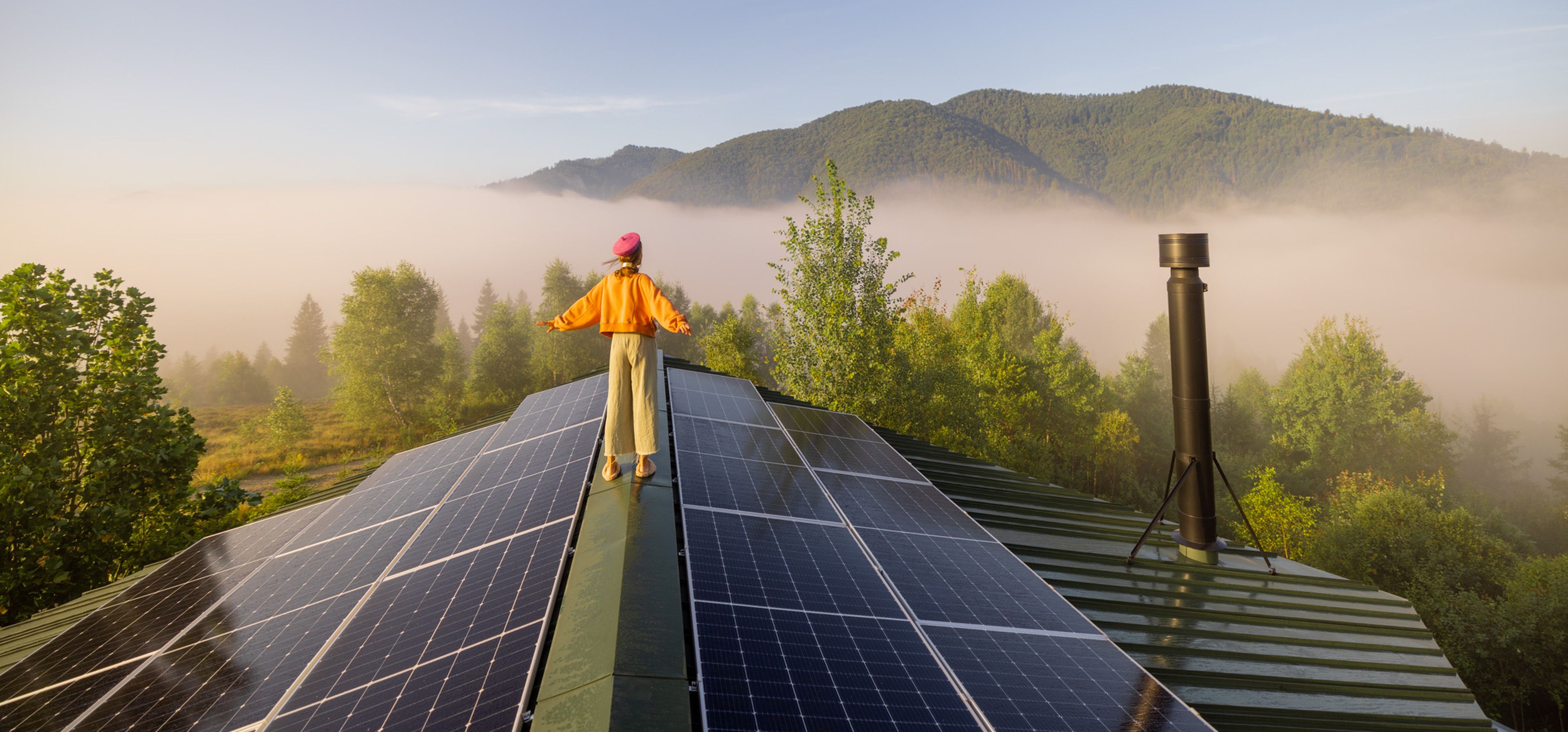 Eine Frau steht mit ausgestreckten Armen auf dem Giebel ihres Hauses und genießt die Sonne & Aussicht. Rechts und links von ihr befinden sich Solarpanelen.
