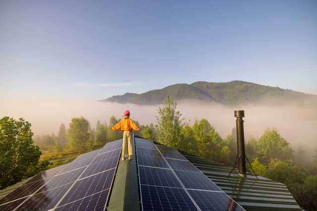 Eine Frau steht mit ausgestreckten Armen auf dem Giebel ihres Hauses und genießt die Sonne & Aussicht. Rechts und links von ihr befinden sich Solarpanelen.