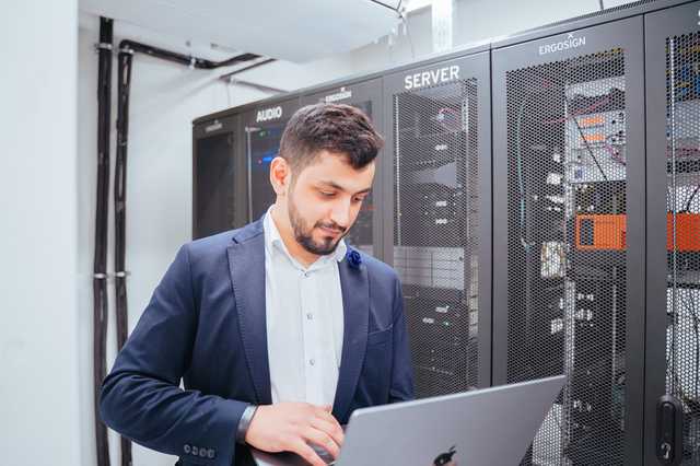 Mann steht mit Laptop vor Servern.