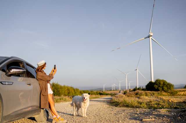Eine Frau lehnt sich an ein Auto zur linken Seite des Bildes und fotografiert Windräder vor einem blauen Himmel. Ein weißer Hund steht neben ihr.