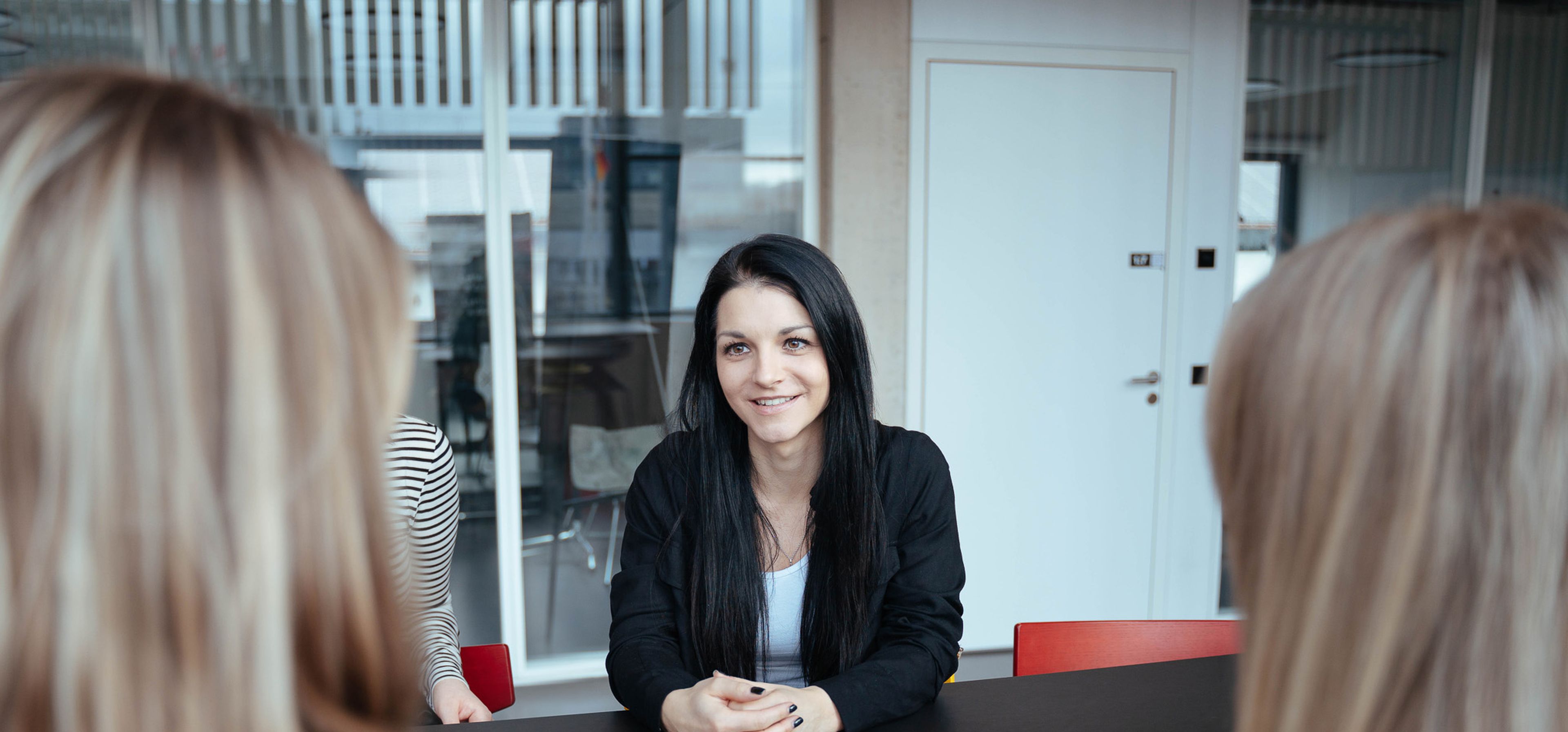Vier Frauen sitzen am Tisch. Das Foto wurde über die Schulter von zwei blonden Frauen aufgenommen. Im Fokus ist eine Frau mit schwarzen langen Haaren auf der gegenüberliegenden Seite und ein Teil der Schulter der Frau neben dieser.