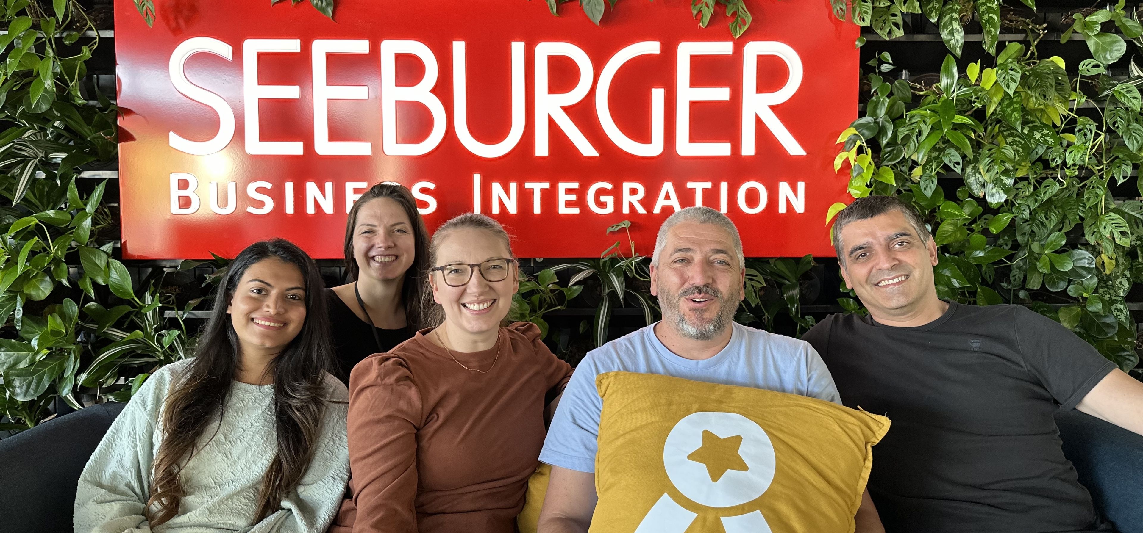 Das Seeburger-Team und eine Mitarbeiterin von Ergosign sitzen gemeinsam auf der Couch. Hinter ihnen hängt das Seeburger-Logo an der Wand.