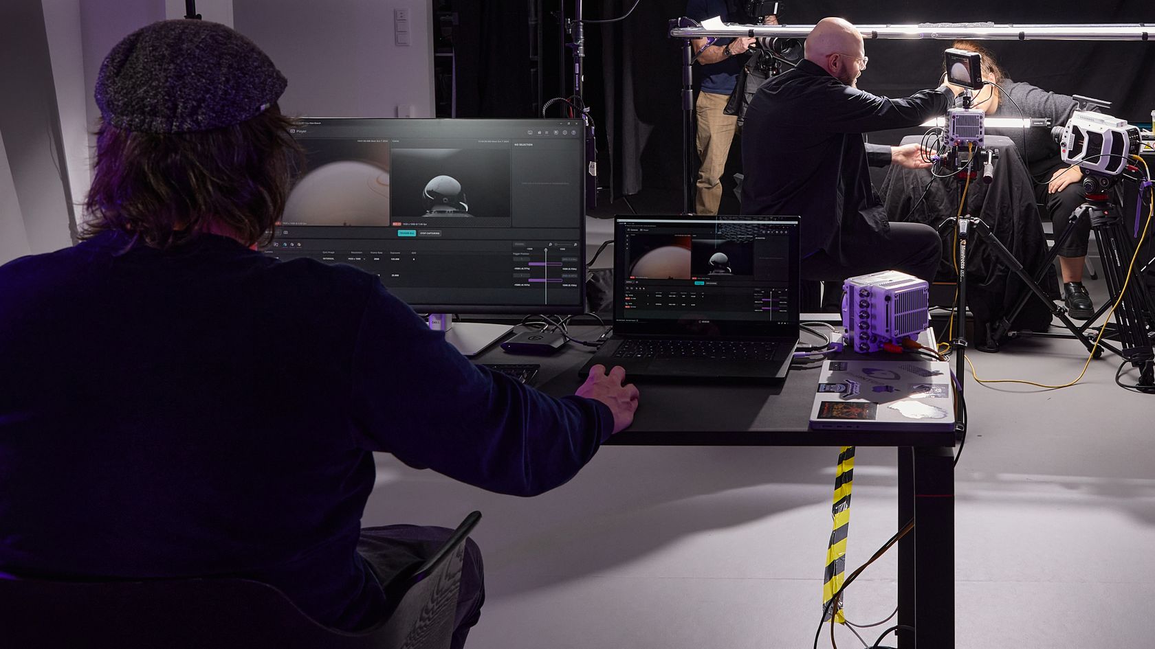 A man sitting at a desk with 2 screens in front of him, on which the Vision Research application can be seen. In the background you can see a photo setting with the high-speed cameras