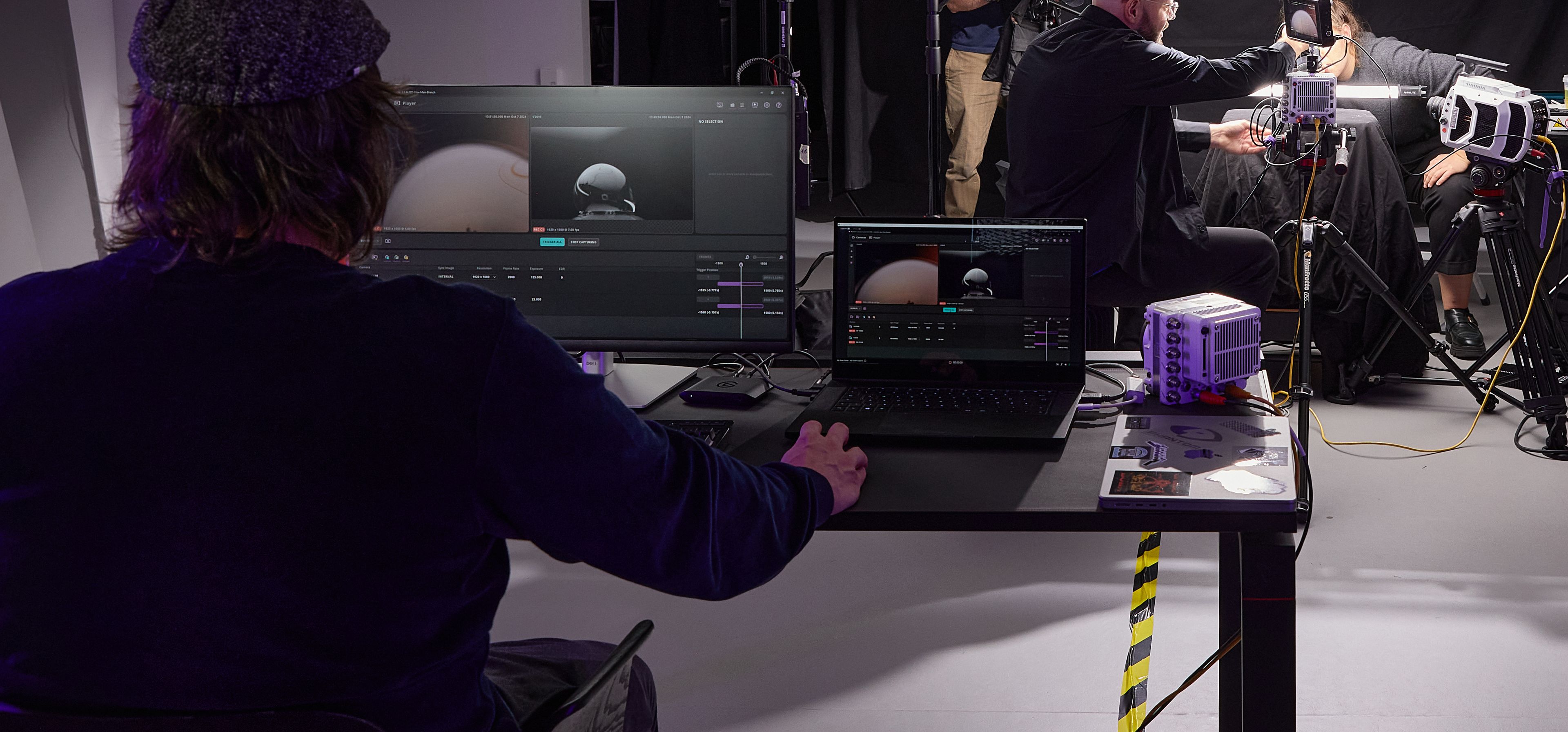 A man sitting at a desk with 2 screens in front of him, on which the Vision Research application can be seen. In the background you can see a photo setting with the high-speed cameras