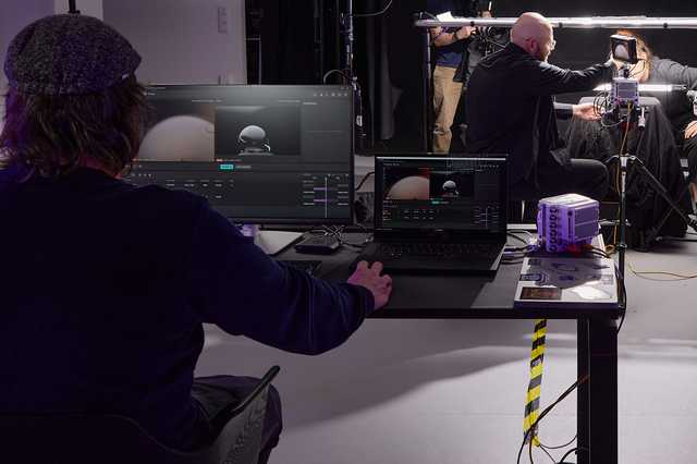 A man sitting at a desk with 2 screens in front of him, on which the Vision Research application can be seen. In the background you can see a photo setting with the high-speed cameras