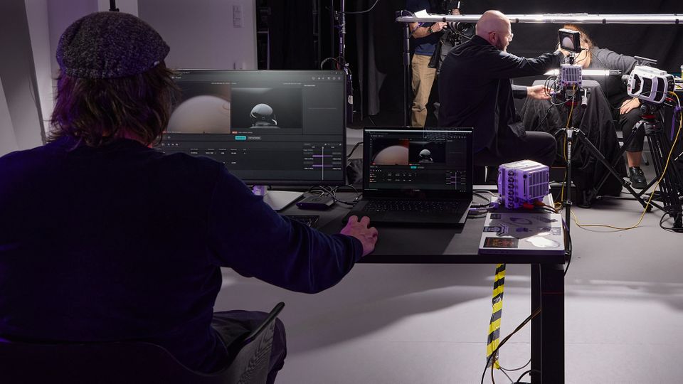 A man sitting at a desk with 2 screens in front of him, on which the Vision Research application can be seen. In the background you can see a photo setting with the high-speed cameras