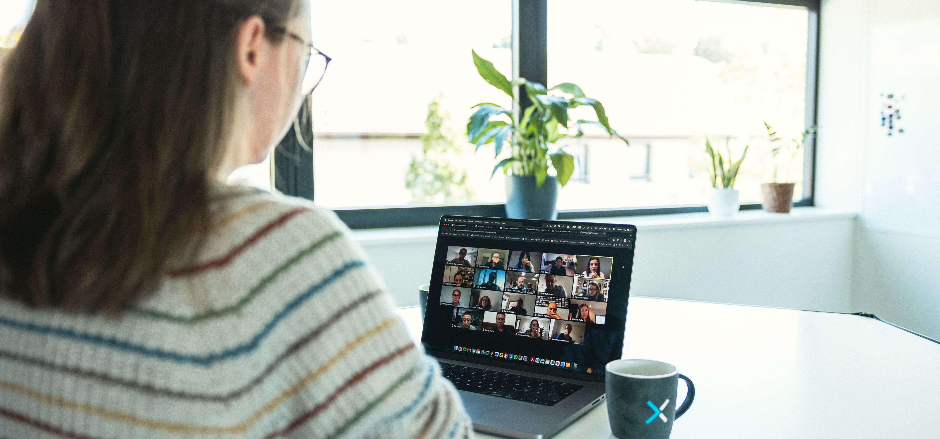Frau im gestrickten Pullover nimmt an einer Videokonferenz auf einem Laptop teil, mit einer Tasse und Pflanzen im Hintergrund.