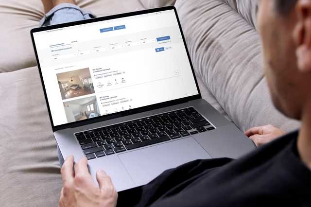 Person auf Sofa durchsucht mit Laptop Immobilienangebote auf einer Webseite. Bildschirm zeigt Wohnungen mit Details und Fotos.
