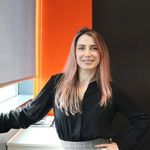 A woman with long pink hair stands smiling by a window in an office, wearing a black blouse and plaid skirt. The background features an orange and black wall.