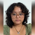 A person with curly hair wearing glasses, a green top, and a butterfly necklace against a white background.