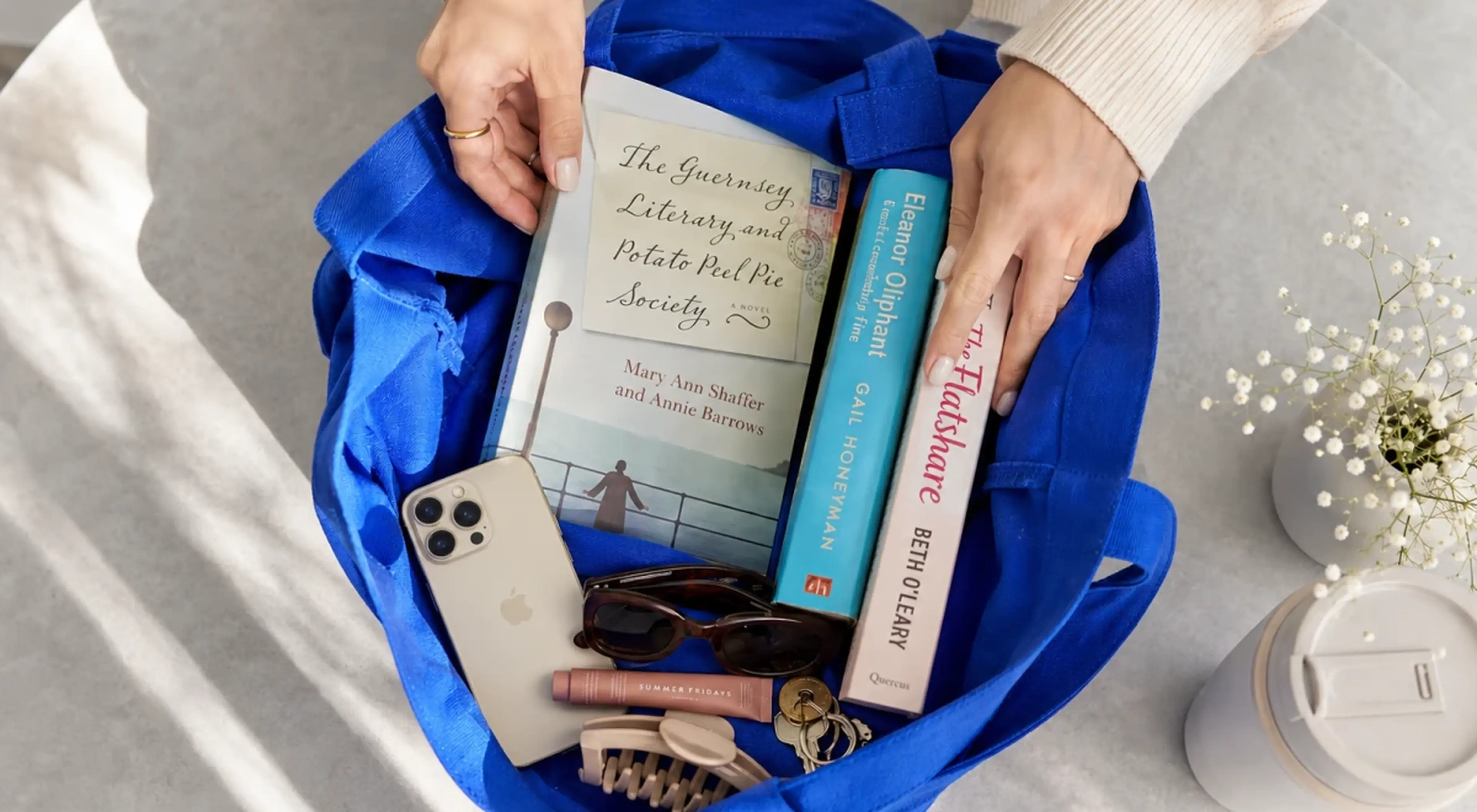 Woman placing feel-good books including The Guernsey Literary and Potato Peel Pie Society, Eleanor Oliphant is Completely Fine, and The Flatshare into a blue tote bag with a phone, sunglasses, and key