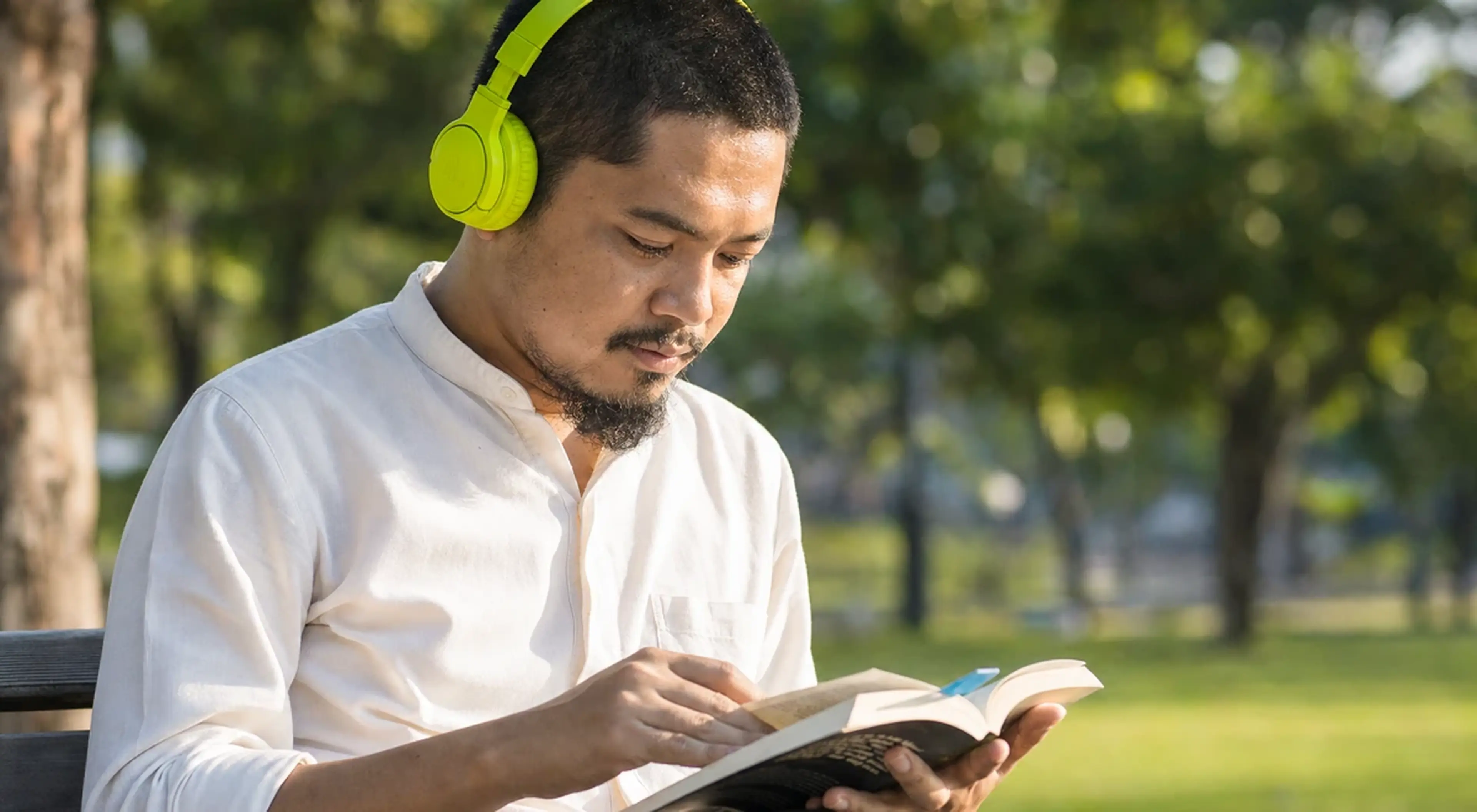 Man wearing green headphones sitting on a park bench, focused on intensive reading of a book surrounded by trees and greenery