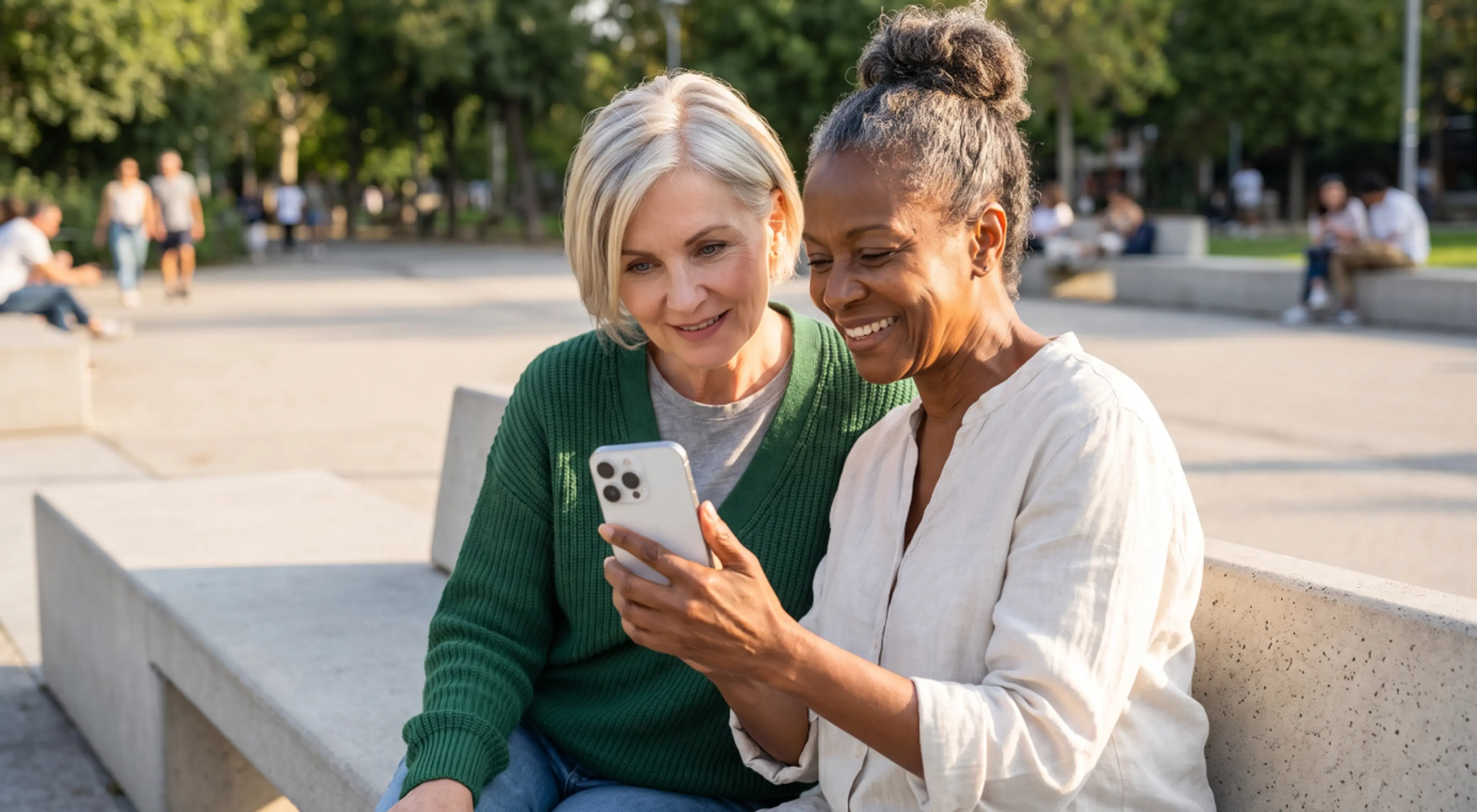 Two smiling mature women sitting on a stone bench in a sunny park, looking together at a smartphone while exploring the best brain training apps