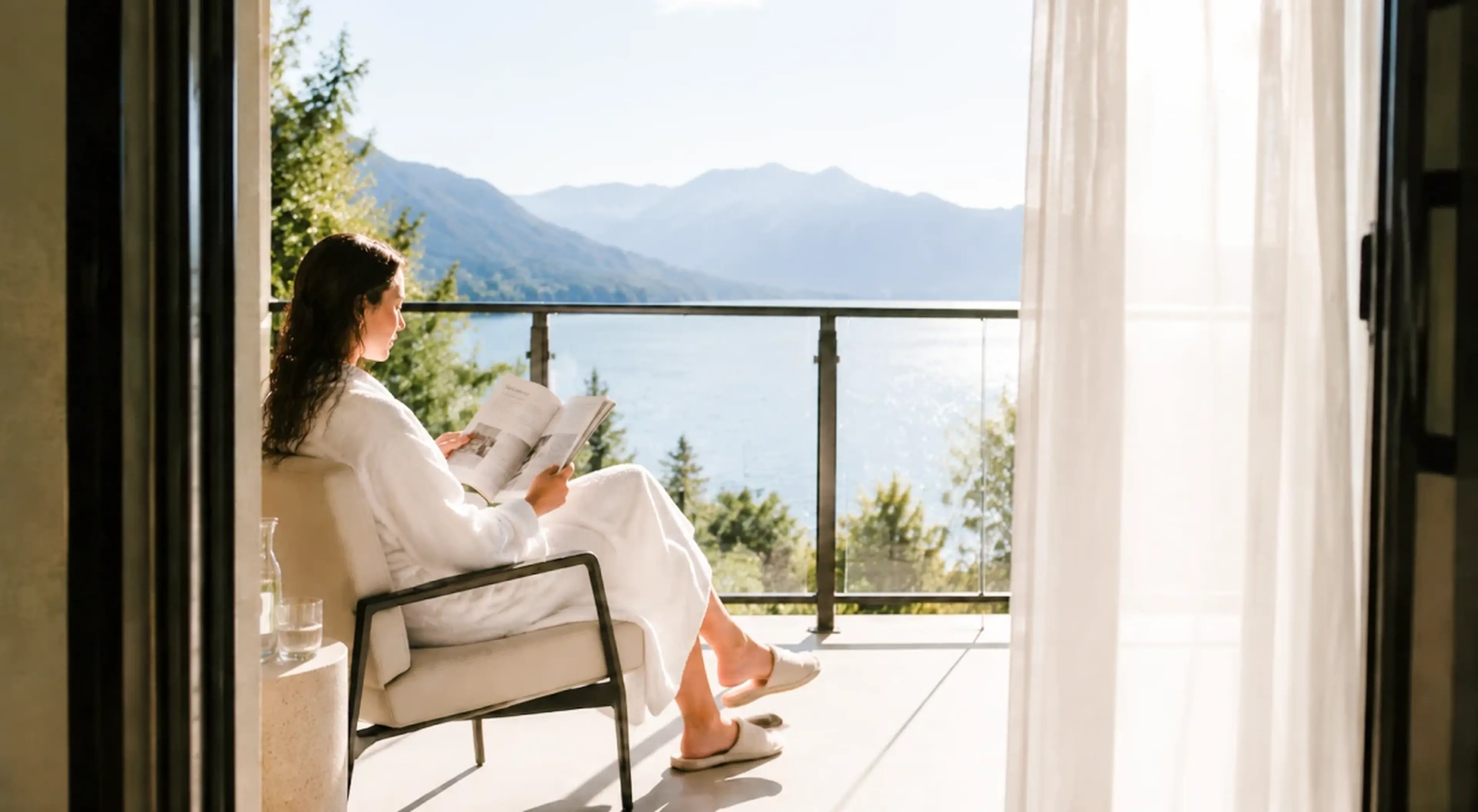 Woman sitting on a sunlit balcony reading, with a mountain lake view and sheer curtains in the background
