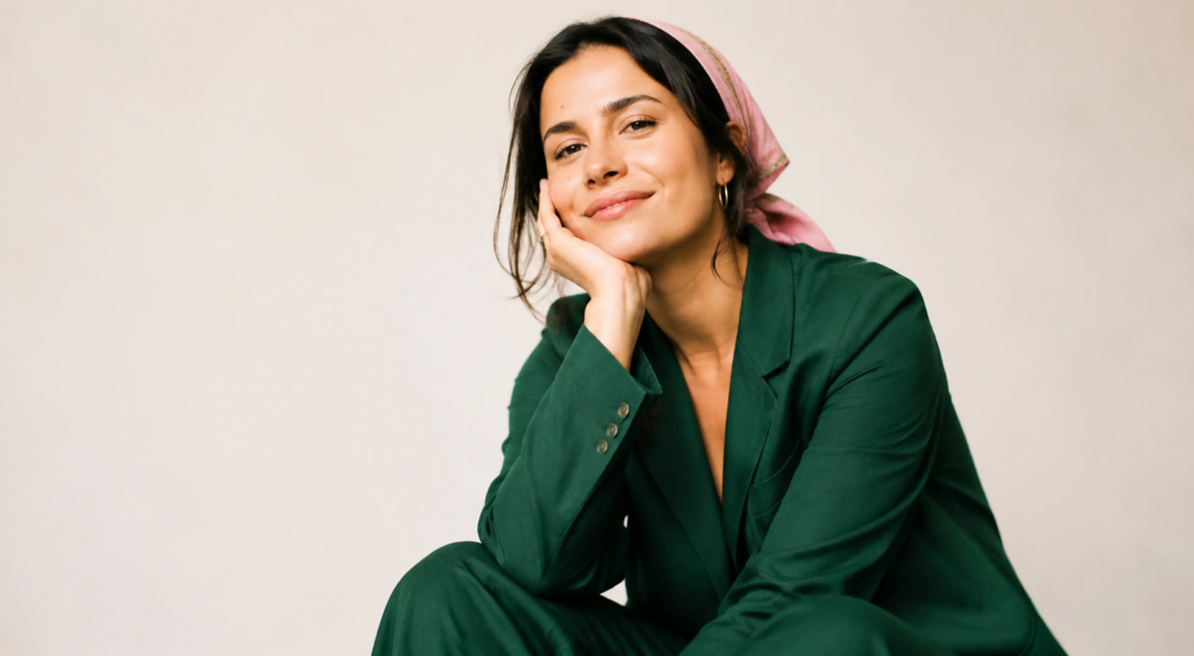 Smiling woman in a green blazer and pink headscarf resting her chin on her hand against a neutral beige backdrop, embodying be the woman he can't forget confidence