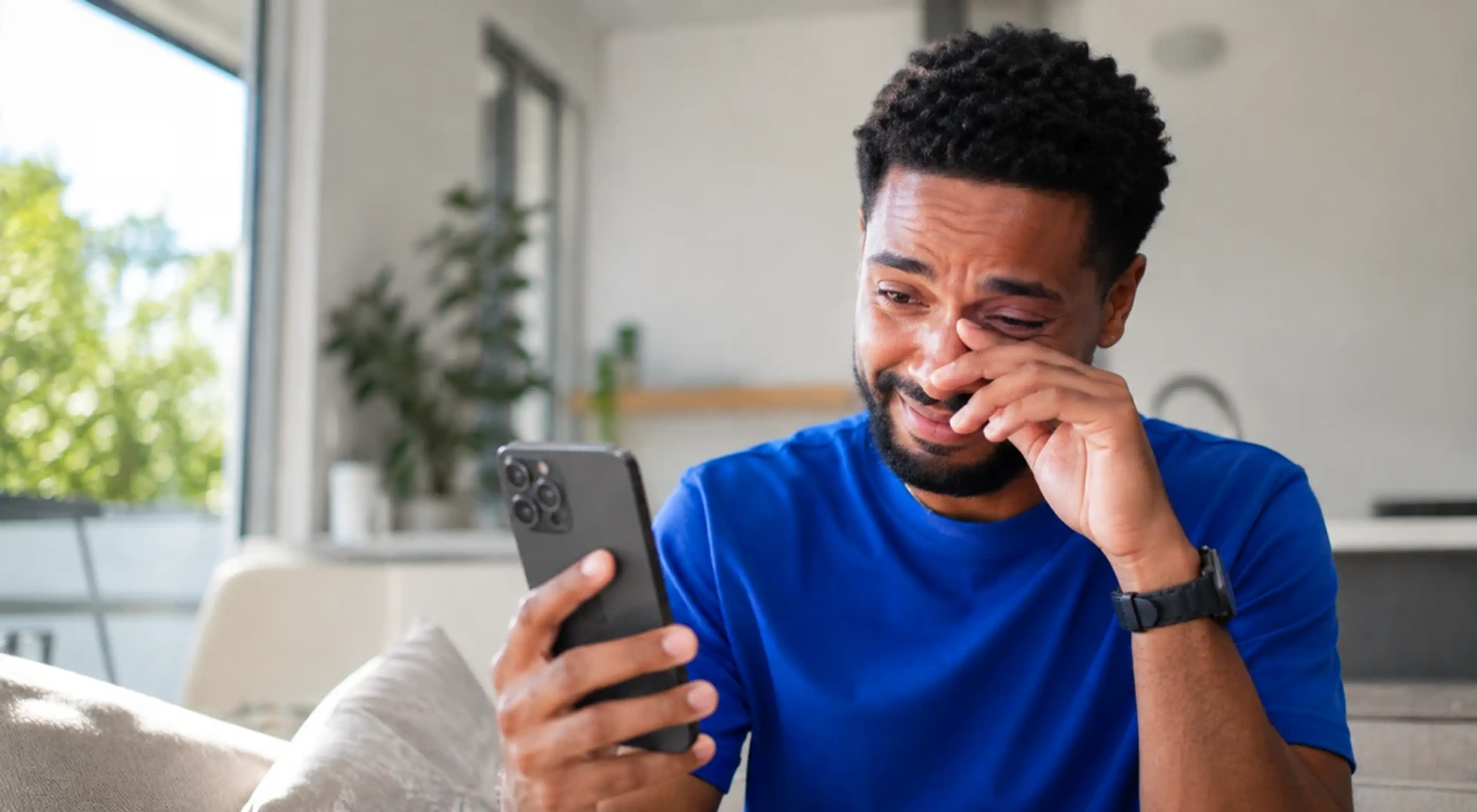 Young Black man in a blue t-shirt crying while looking at his smartphone on a couch at home, showing emotional sensitivity and vulnerability in a bright modern living room