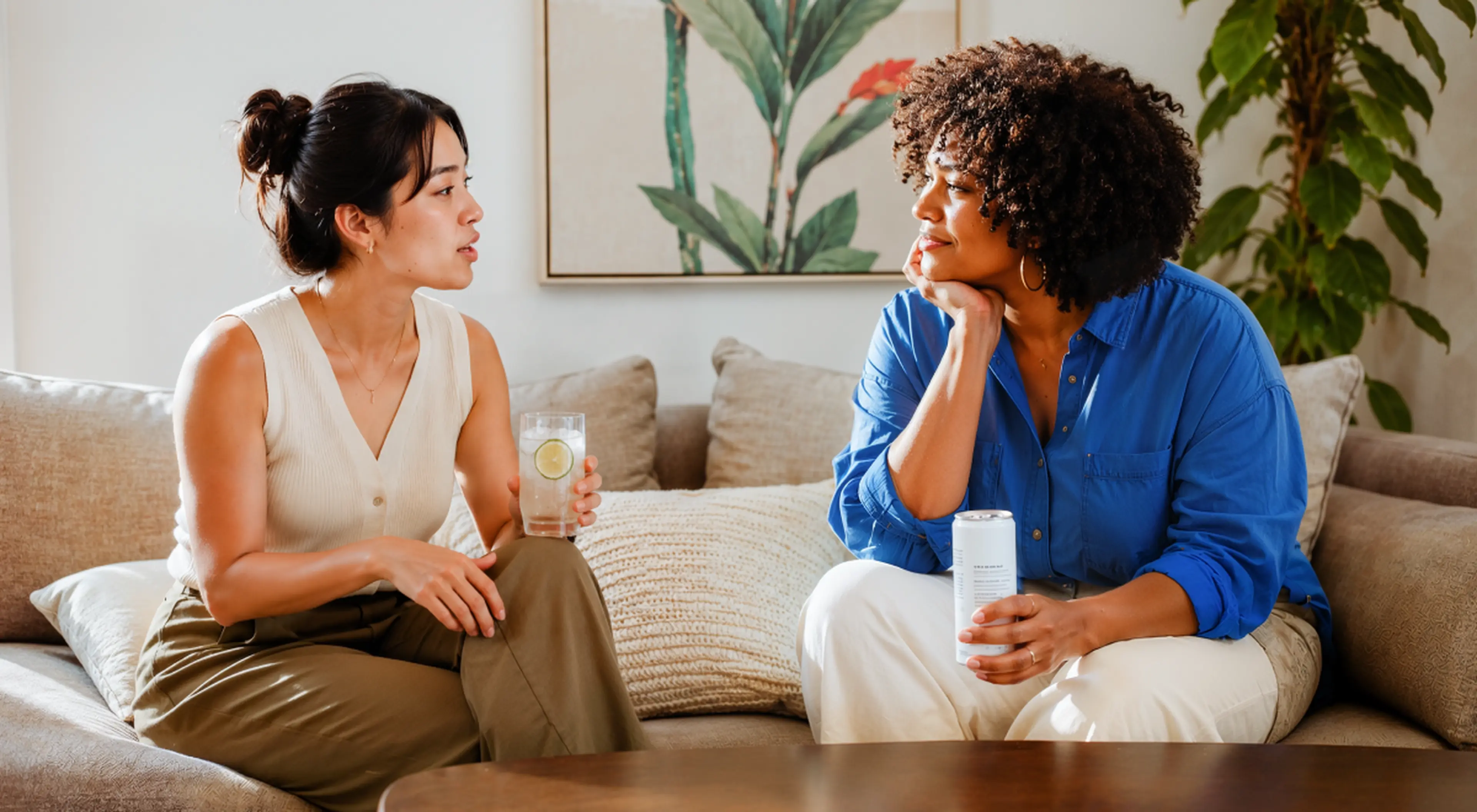 Two women practicing communication skills while talking on a beige sofa, one holding a lemon water glass, the other a ca