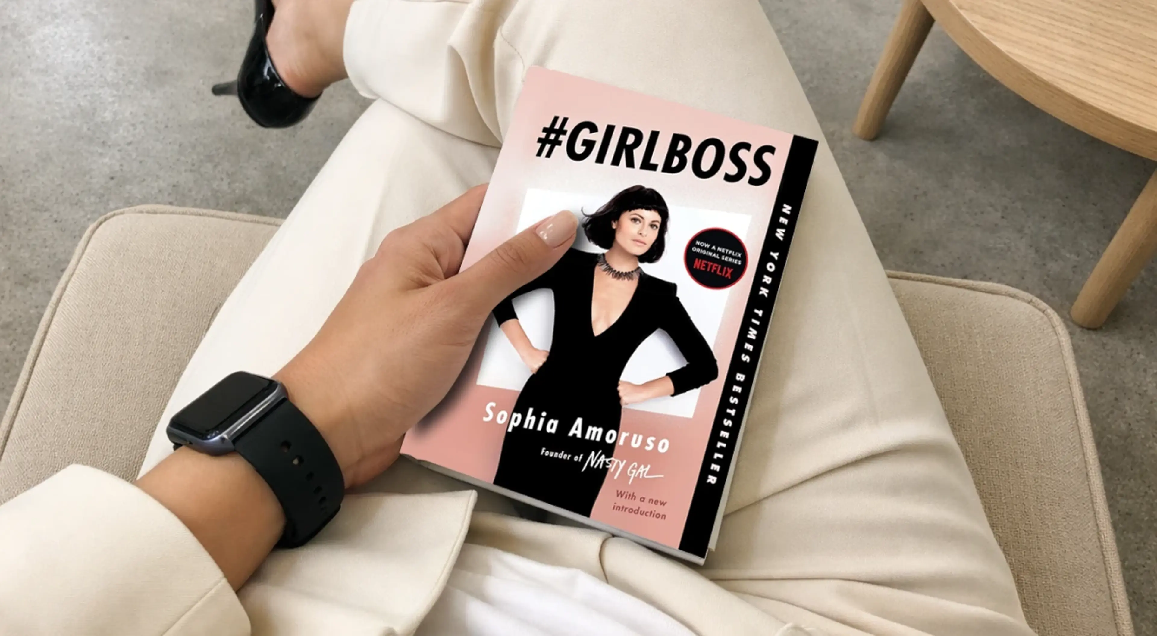 Woman wearing a black Apple Watch holding the #GIRLBOSS book by Sophia Amoruso while sitting on a beige couch, black heels visible in the background — self-growth read for a woman CEO