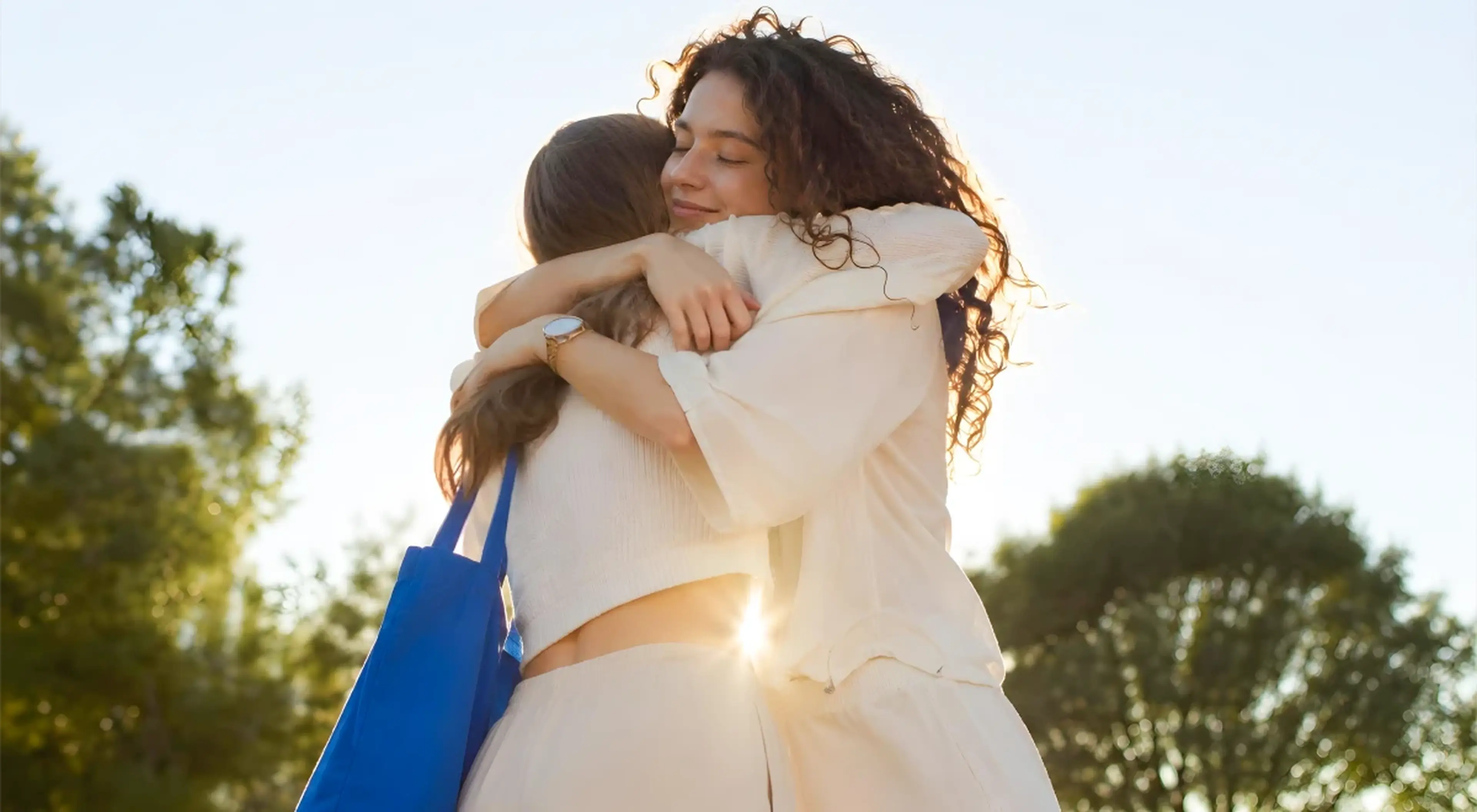 Two women sharing a warm embrace outdoors in sunlight with green trees in the background, representing forgiveness and emotional reconciliation