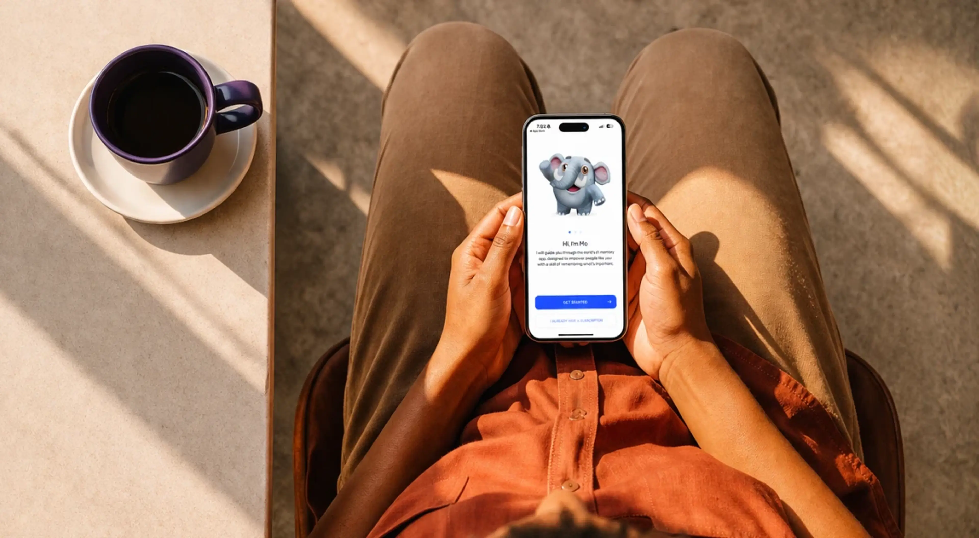 Person sitting cross-legged holding a smartphone with the MemoryOS app open, coffee cup on a saucer nearby, warm natural light setting